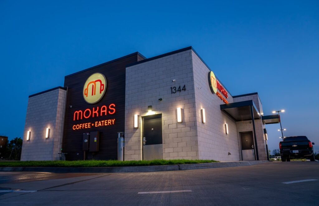 Exterior of a Mokas Coffee + Eatery building at dusk, with the logo and illuminated signage on a modern, light-gray brick facade. Warm wall lights highlight the structure, and the address number ‘1344’ is visible. A pickup truck is parked near the entrance, and the clear evening sky provides a deep blue backdrop